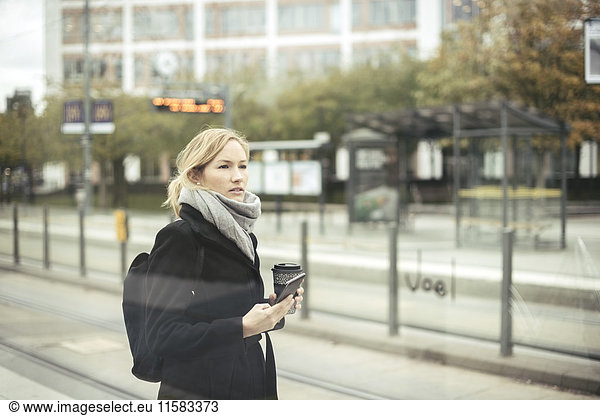 Mid adult businesswoman holding mobile phone and disposable coffee cup at tram station