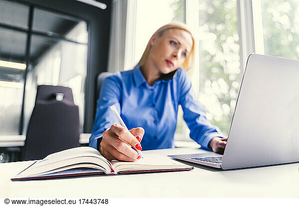 Mid adult businesswoman writing in diary while sitting with laptop at desk in office