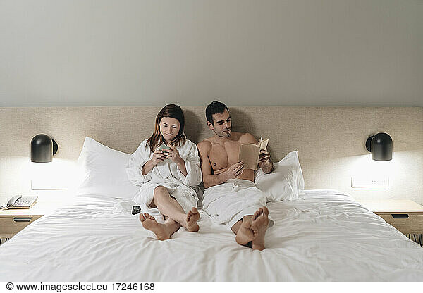 Mid adult couple reading book and using smart phone on bed