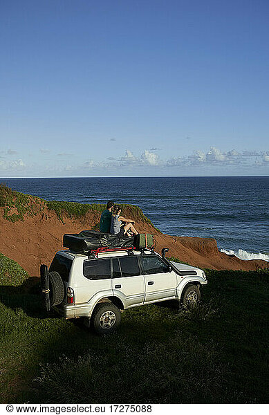 Mid adult couple sitting on 4x4 roof while looking at sea against blue sky