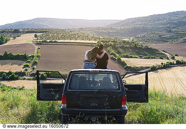 Mid adult couple sitting on 4x4 vehicle roof while looking at landscape during road trip
