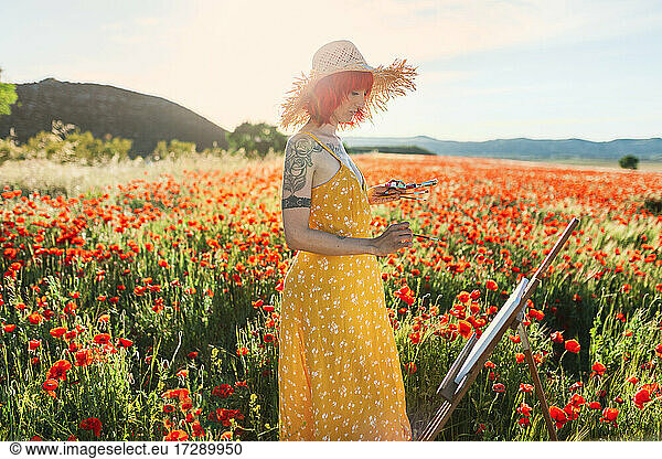 Mid adult female artist in yellow sundress painting on poppy field