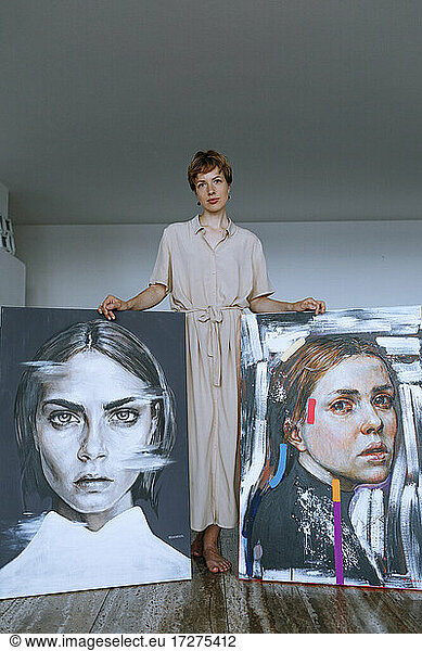 Mid adult female artist standing with paintings against wall in art studio