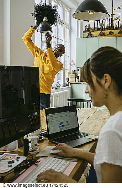 Mid adult man cleaning pendant light while female freelancer using computer working at home