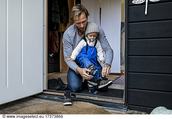Mid adult man helping son put on shoe at doorway