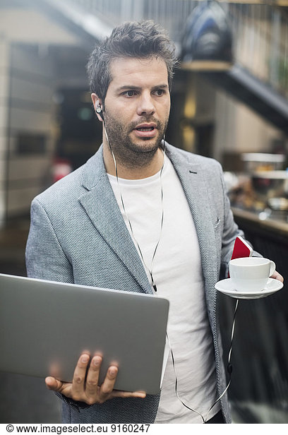Mid adult man holding laptop and coffee in cafe