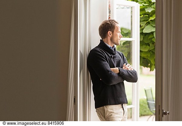 Mid adult man looking out of window  arms folded