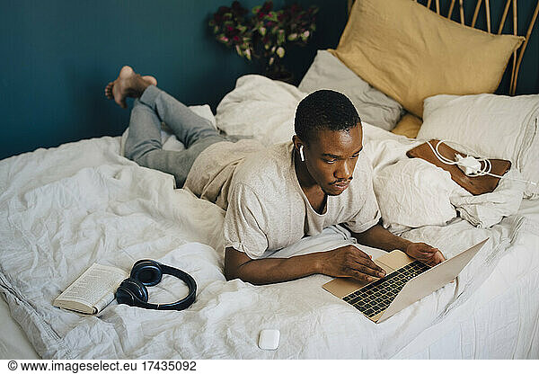 Mid adult man using laptop while wearing wireless in-ear headphones in bedroom