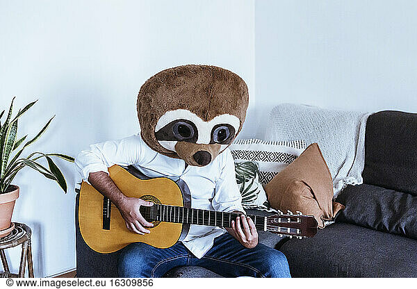 Mid adult man wearing teddy bear mask playing guitar while sitting on sofa in living room