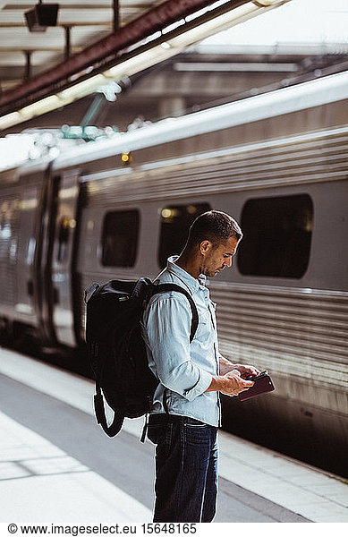Mid adult man with backpack using mobile phone while standing by train on platform