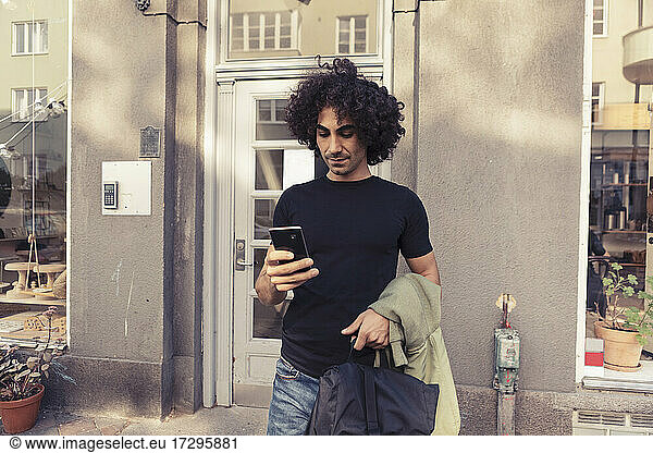 Mid adult man with curly hair using smart phone while standing outside cafe