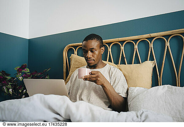 Mid adult man working on laptop while having coffee in bed