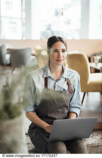 Mid adult manager in apron sitting with laptop while contemplating in coffee shop