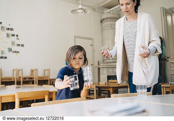 Mid adult teacher with boy arranging drinking glasses on table in classroom at child care