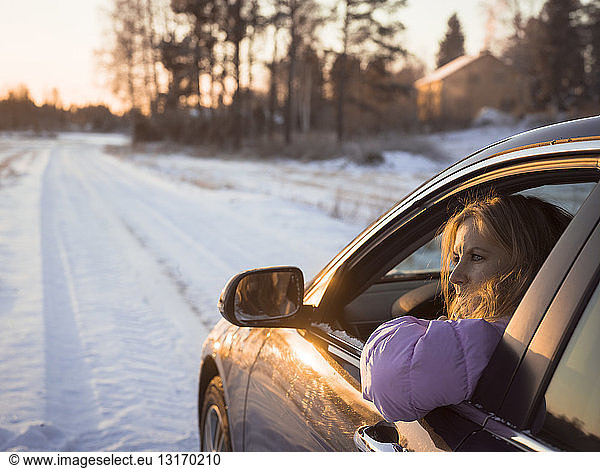 Mid adult woman driving car along snowy road  at sunset