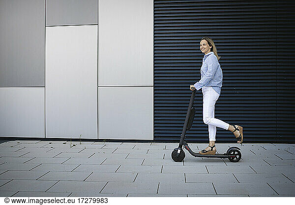 Mid adult woman driving electric push scooter on footpath by metal wall