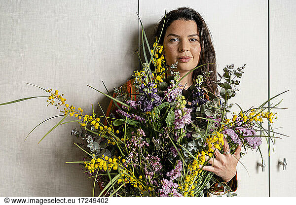 Mid adult woman holding bunch of flowers while standing in front of wall