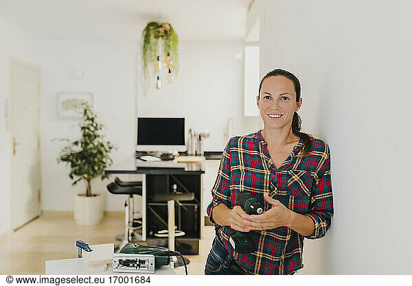 Mid adult woman holding electric drill while leaning on wall at home