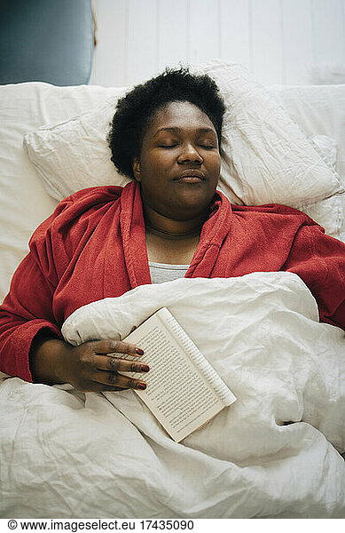 Mid adult woman in red bathrobe sleeping with book on bed at home