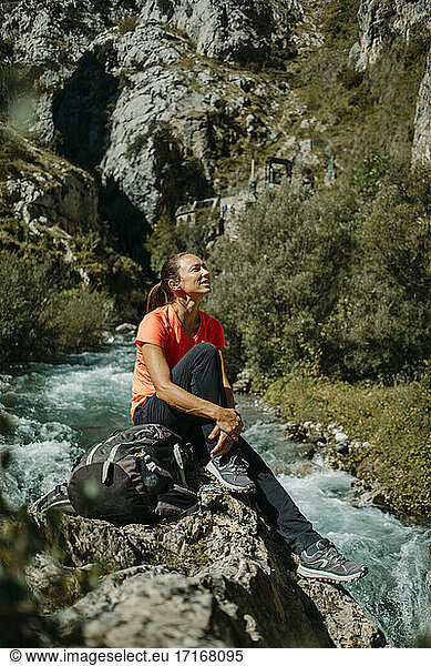 Mid adult woman looking away while sitting by river at Cares Trail in Picos De Europe National Park  Asturias  Spain