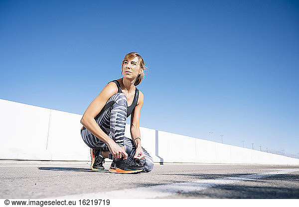 Mid adult woman looking away while tying shoelace on road against clear blue sky