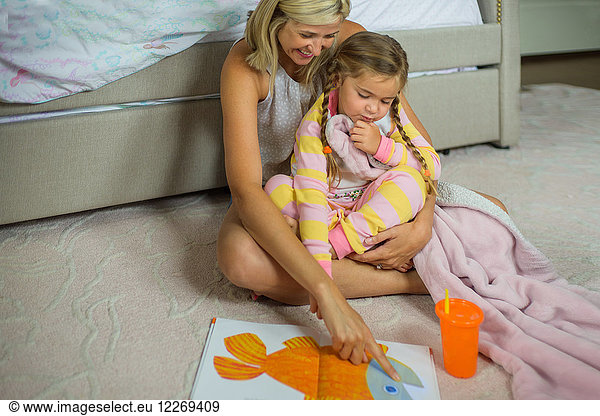 Mid adult woman sitting on floor with daughter at bedtime looking at picture book