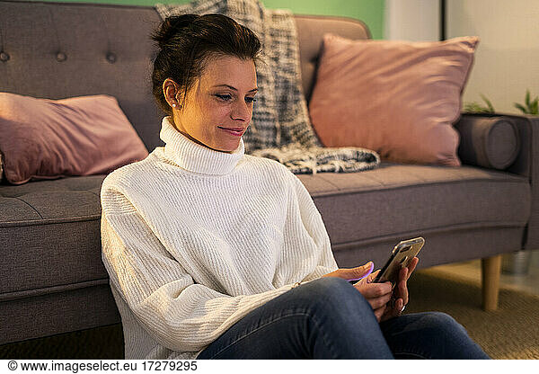 Mid adult woman text messaging on smart phone while sitting against sofa at home