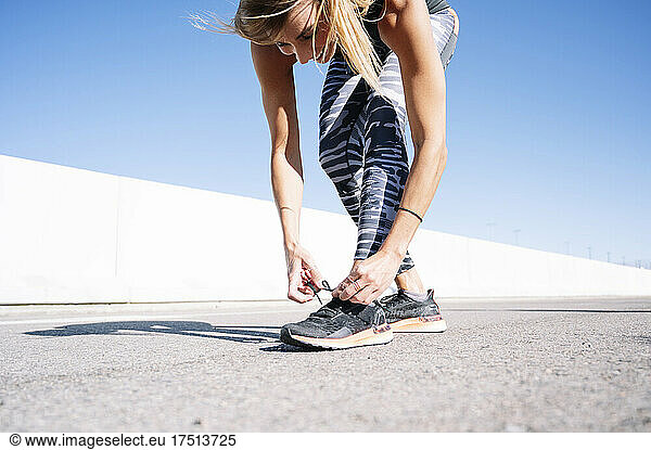 Mid adult woman tying shoelace on road against clear blue sky in city