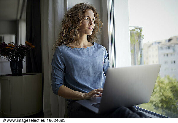 Mid adult woman using laptop while sitting on window sill at home