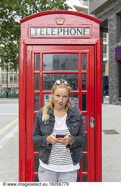Mid adult woman using smart phone while standing against telephone booth