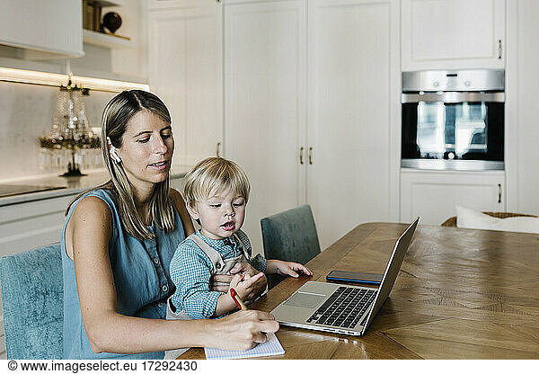 Mid adult woman writing on diary during video call while son sitting on lap sitting at home