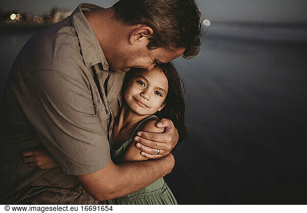 Mid-40's dad hugging 8 yr old daughter on beach at sunset