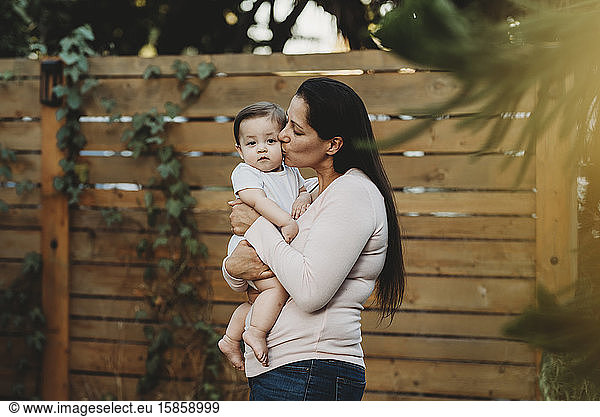 Mid-30's mom holding and kissing baby girl in backyard near fence