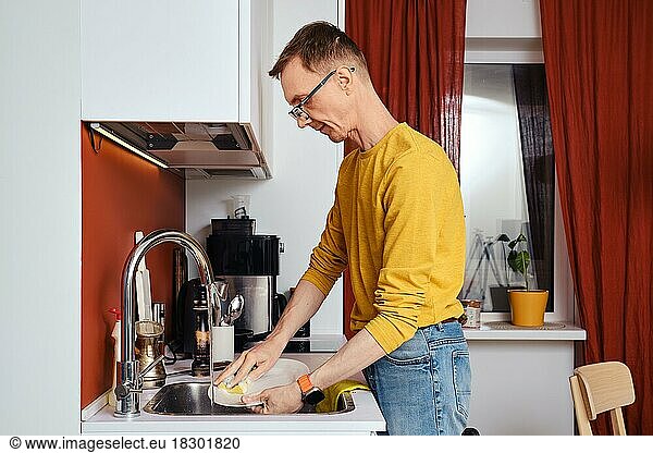 Middle-aged man in spectacles washing dishes in kitchen in the evening