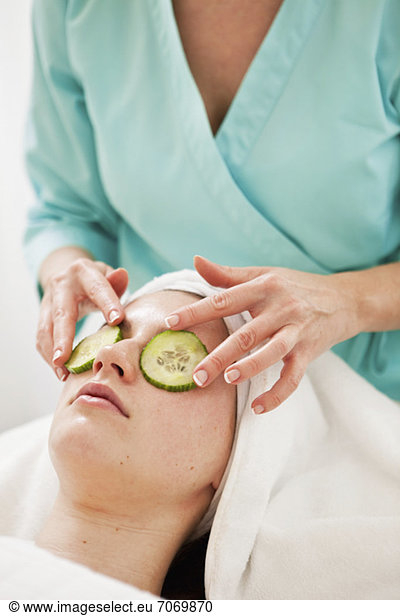 Midsection of a beautician keeping cucumber slices on young woman' eyes in beauty spa