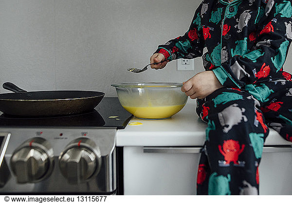Midsection of boy mixing batter in bowl while sitting on kitchen counter at home
