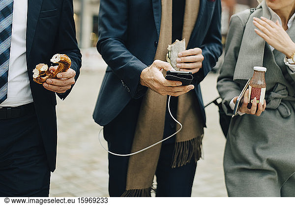 Midsection of business professionals holding pretzel and drink while using smart phone in city