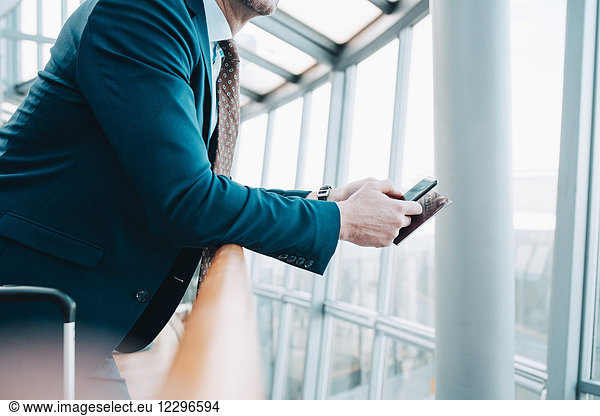 Midsection of businessman holding mobile phone and passport while leaning on railing at airport