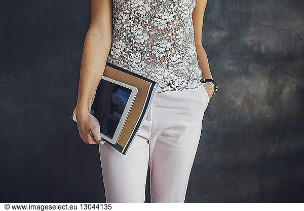 Midsection of businesswoman holding tablet and files while standing against wall at home office