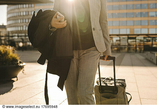 Midsection of businesswoman with smart phone pulling luggage while walking on sunny day