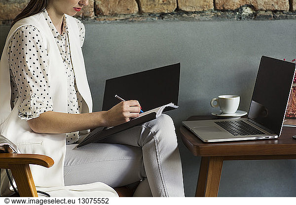 Midsection of businesswoman writing in file with laptop at table in hotel lobby