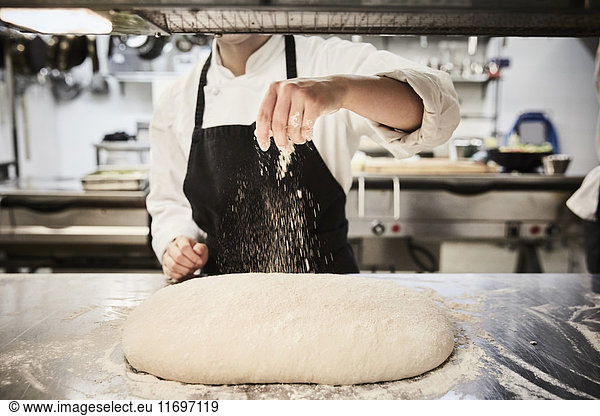 Midsection of female chef student sprinkling flour on dough at commercial kitchen