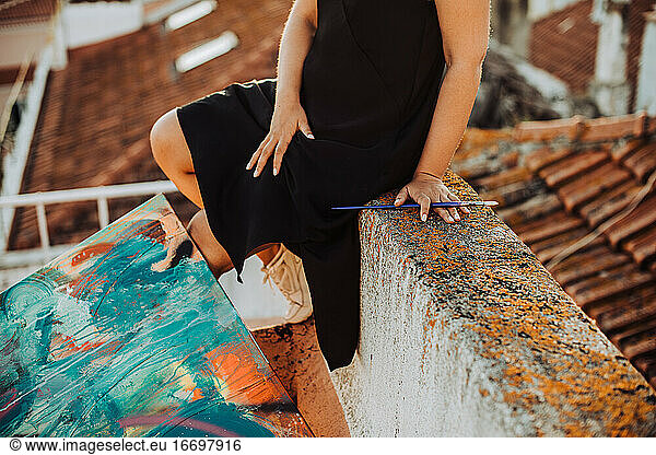 Midsection of female painter sitting on roof with abstract painting