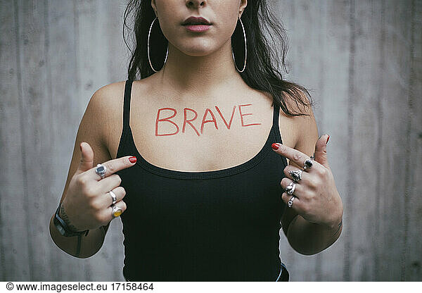 Midsection of female protestor pointing on brave text written over chest against wall