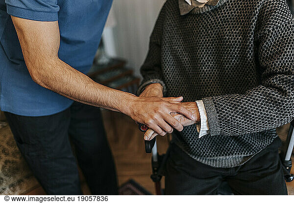Midsection of male care assistant holding hands of senior man at home