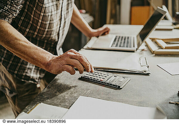 Midsection of male carpenter using calculator on workbench at repair shop