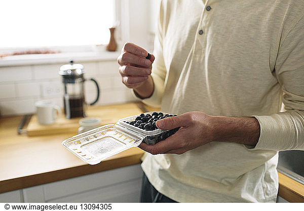 Midsection of man holding box with blueberries while leaning on kitchen counter at home