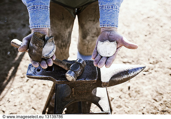 Midsection of man holding hoof while standing by hammer and anvil on field