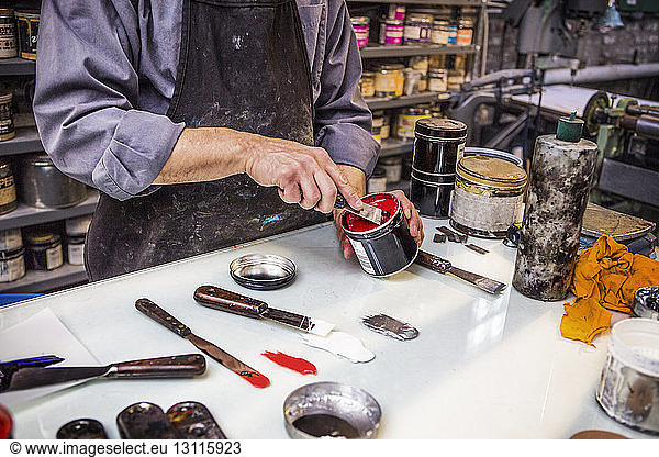 Midsection of man removing paint from can with ink knife on table