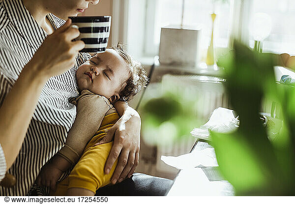 Midsection of mother drinking coffee while carrying sleeping baby boy at home